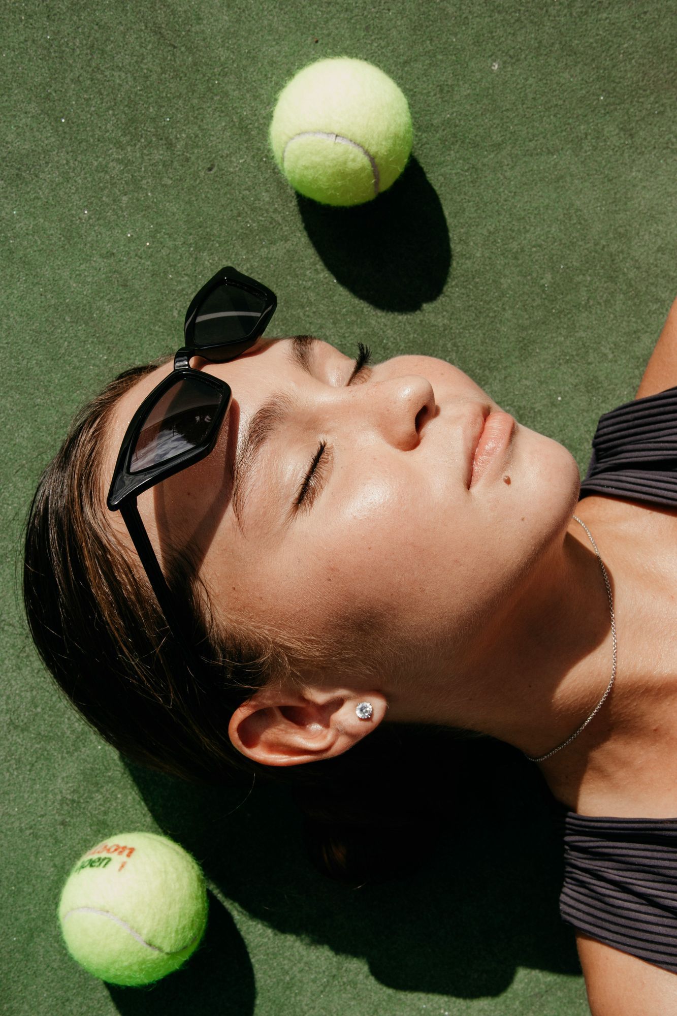 Woman lying on a tennis court with eyes closed, wearing sunglasses and a dark tank top, surrounded by tennis balls—capturing a moment of rest and sunlight after play.