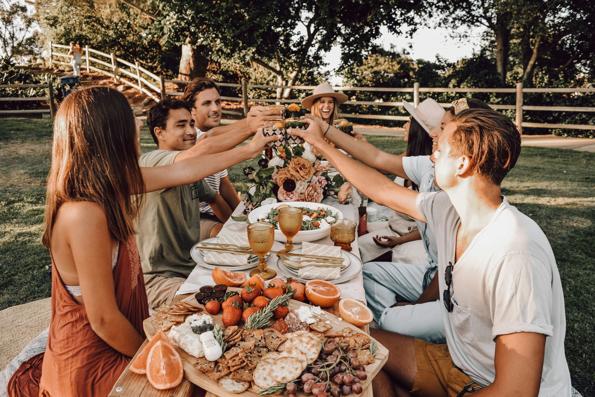 Group of friends gathered around an outdoor table at VERVE Columbia, toasting with drinks over a spread of fresh fruit, cheeses, and snacks in a sunny, relaxed setting.