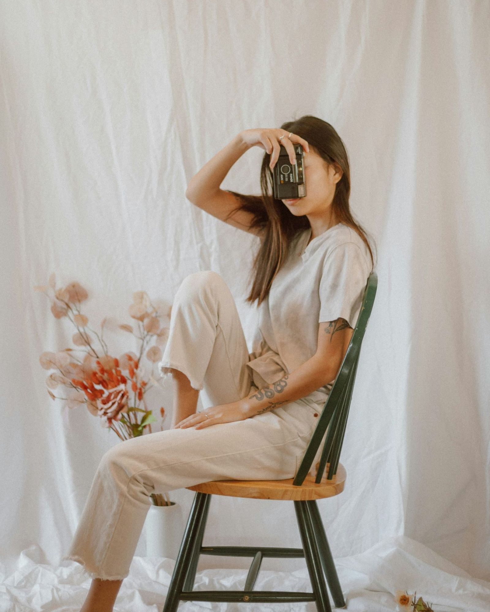 A woman sitting on a chair holding a camera up to her face, dressed in neutral tones with a soft backdrop and floral arrangement—capturing the creative lifestyle vibe of VERVE Columbia.