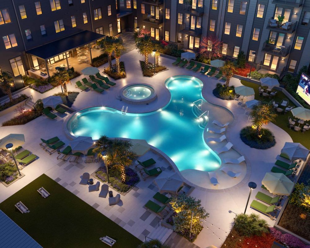 Aerial view of the illuminated pool courtyard at VERVE Columbia at night, featuring glowing water, palm trees, and ample lounge seating in a vibrant, resort-style setting.